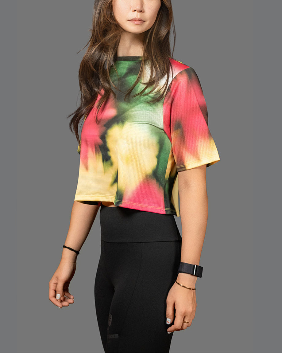 A woman modeling a short-sleeve crop top with a vibrant tie-dye pattern in red, green, yellow, and orange, paired with black leggings, against a solid gray background.